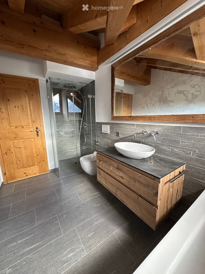 Bathroom with gray tiles, a wooden door, a large mirror, a white sink, a wooden cabinet, a glass shower, and a white bathtub.