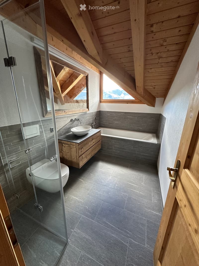 A modern bathroom with wooden fixtures, a glass-enclosed shower, a wooden vanity with a sink, a bathtub, and a toilet. It also features wooden beams and two windows offering a view of the mountains.