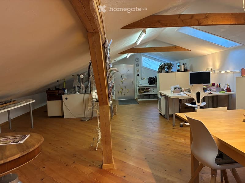 Open layout with wooden floors, beams, and ceiling lights, desks with chairs, computer monitors, a round wooden table, and magazines