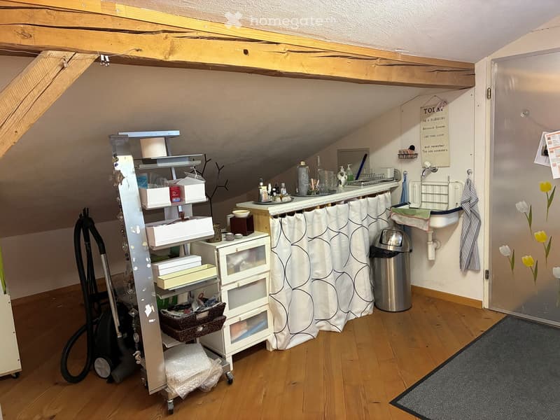 Small attic room, sloped ceiling, wooden floor, kitchenette, storage shelves, black vacuum, trash bin, curtain