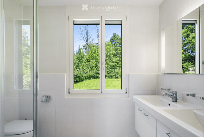 A spacious modern bathroom with white tiles, a glass shower, and a double vanity. The double vanity has two sinks and a mirror with shelves. The room has a window offering a view of a green lawn.