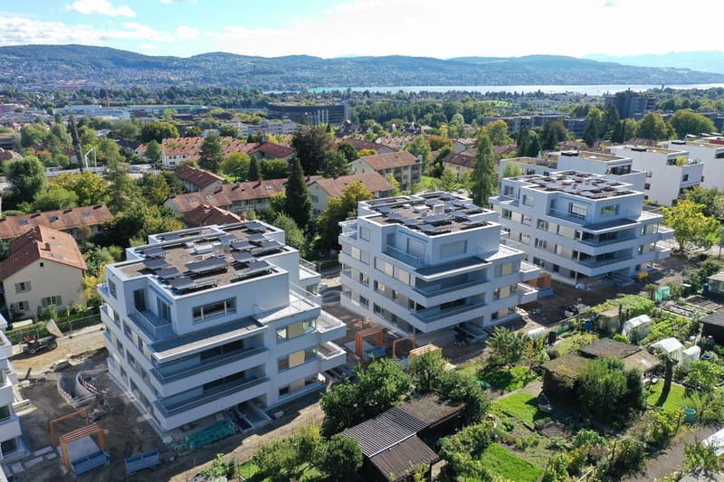 Aerial view of an apartment complex with modern architecture, large windows, balconies, solar panels on the roofs, surrounded by greenery and buildings.
