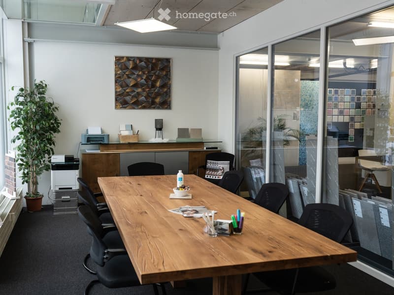 A meeting room with a wooden table, black chairs, glass walls, and a white ceiling. There is a plant, a printer, and a framed painting on the wall. In the distance, another room is visible through the glass wall, containing a desk, a chair, and a blue wal