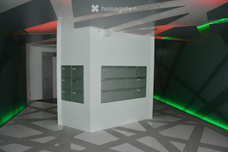 Modern hallway with white walls, geometric ceiling design, and mailboxes on a white pillar. The floor features a black and white geometric pattern, and the lighting includes green neon accents along the walls.