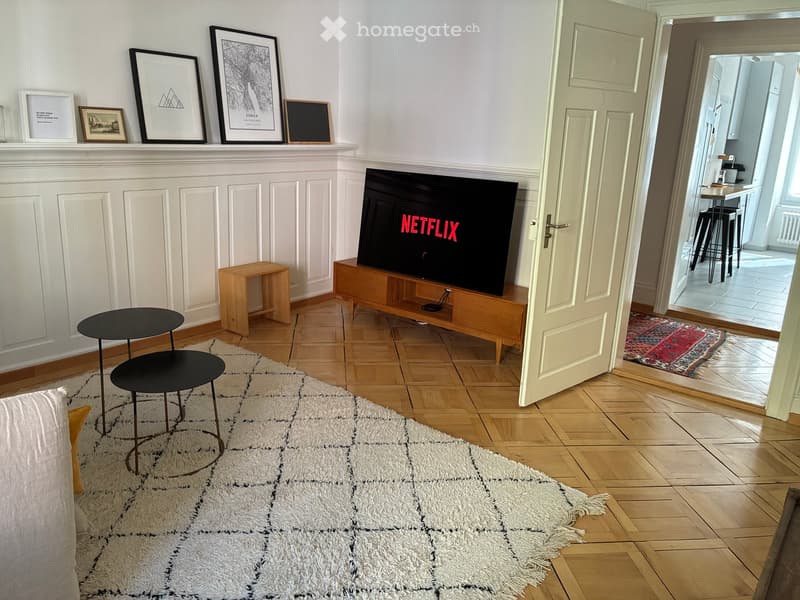 Living room with a fireplace, television displaying Netflix, two black round tables, a wooden stool, and several picture frames on the mantel.