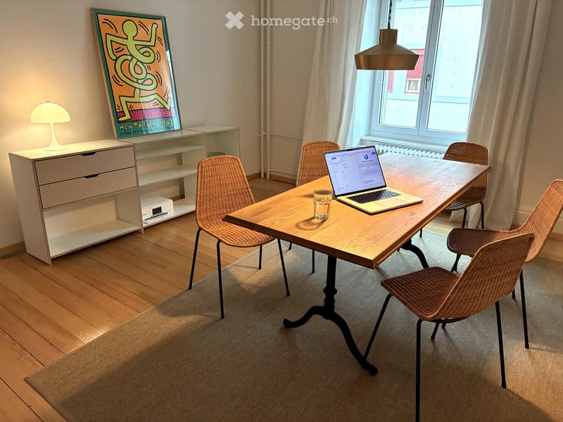 A room with a wooden desk, laptop, glass, and chairs. A lamp is on the cabinet, and a painting is on the wall. A radiator is on the wall near the window.