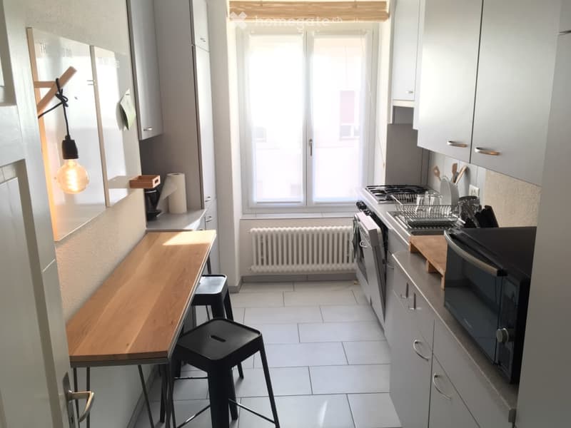 modern kitchen with table, two stools, cabinets, sink, white tiled floor, large window, gas stove, oven, refrigerator, dishwasher, lamp, radiator