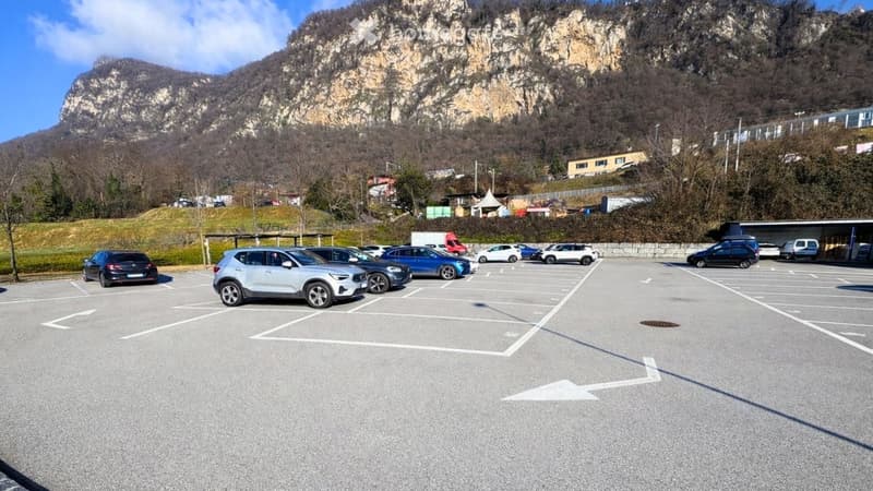 Outdoor parking lot with multiple vehicles parked in rows, a mountain in the background, a building to the right.