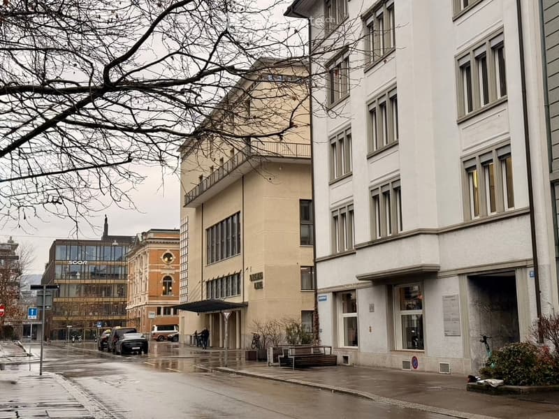 A building with a balcony and arched windows, next to a building with large windows, on a rainy street with cars and pedestrians.