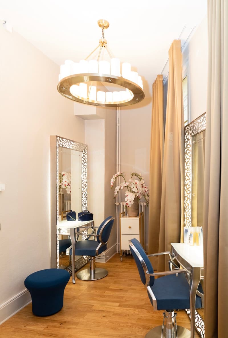 A beauty salon with blue chairs, a large mirror, a table with flowers, and a chandelier.
