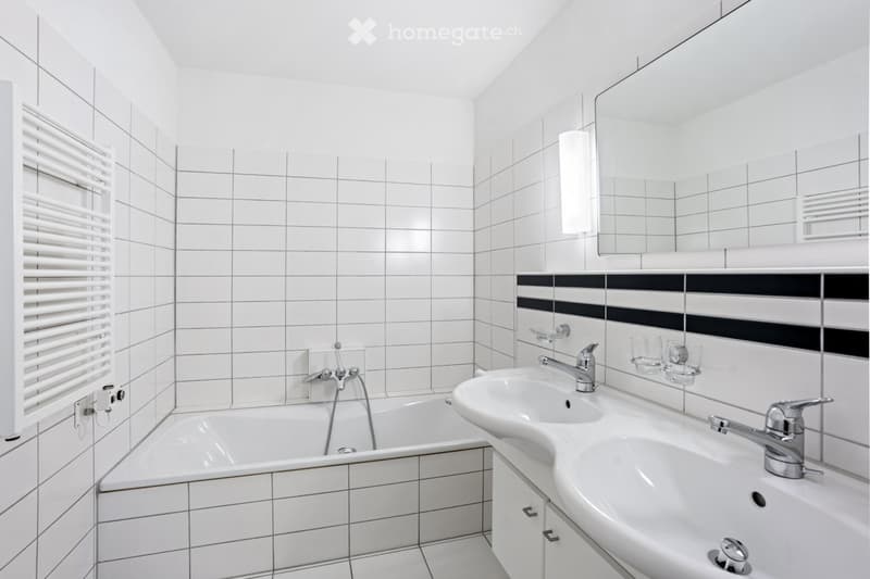 Modern bathroom with white tiled walls and floor, featuring a bathtub, double sink, large mirror, and radiator.
