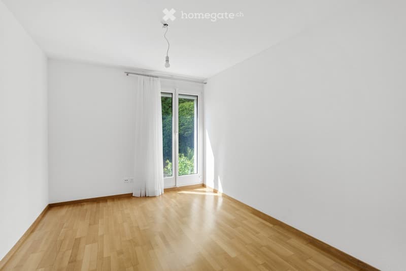 Empty room with white walls, wooden floor, large window with white curtains, and a single light fixture on the ceiling.