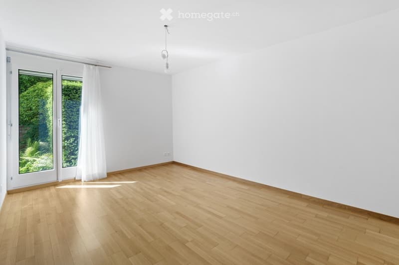Empty room with light wooden flooring, white walls, large windows with white curtains, and a single hanging light fixture.