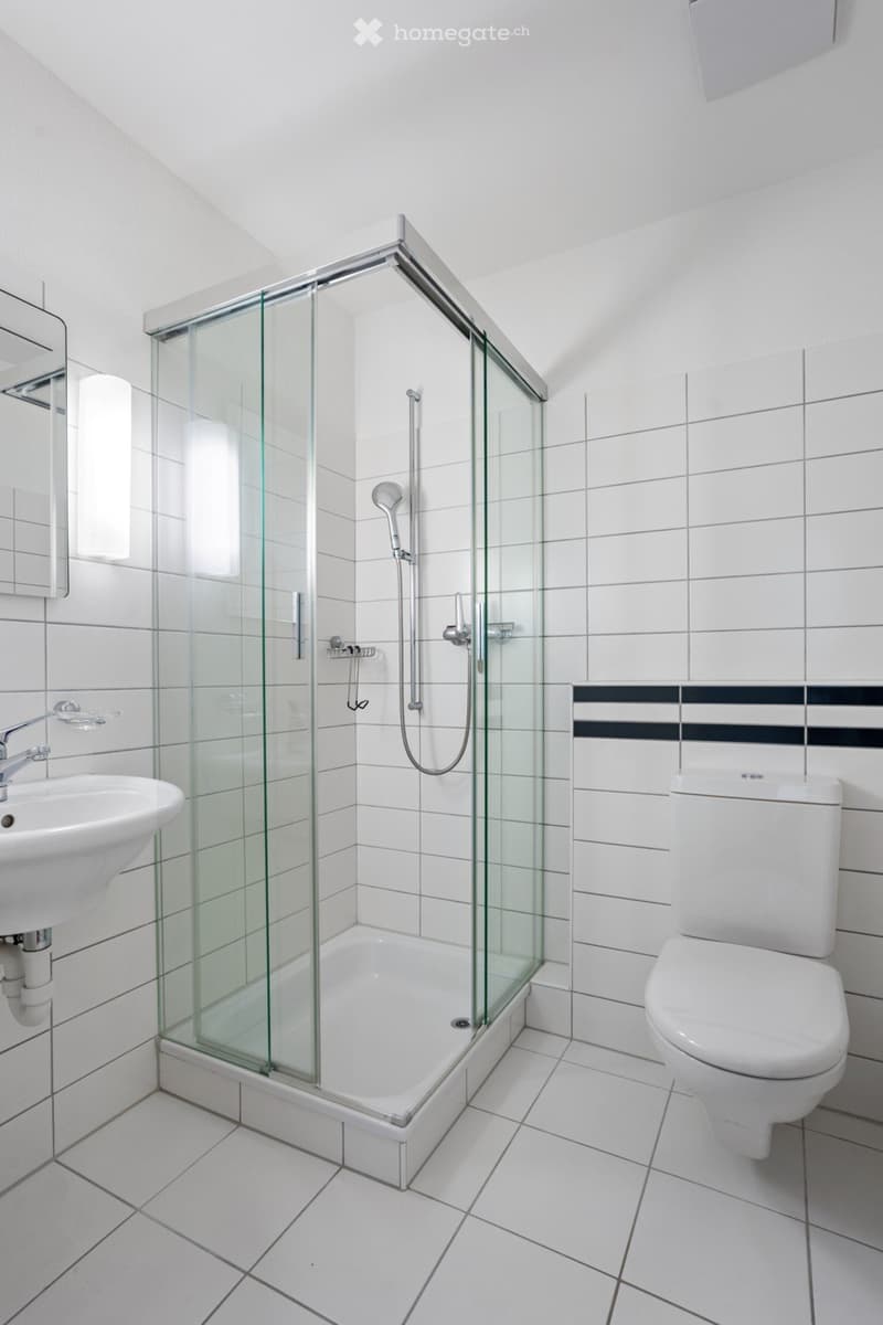Modern bathroom with white tiles, a glass-enclosed shower, a sink, and a toilet.
