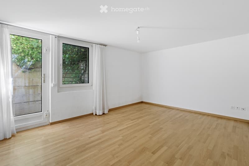 Empty room with wooden flooring, white walls, large windows with white curtains, and a door leading outside.