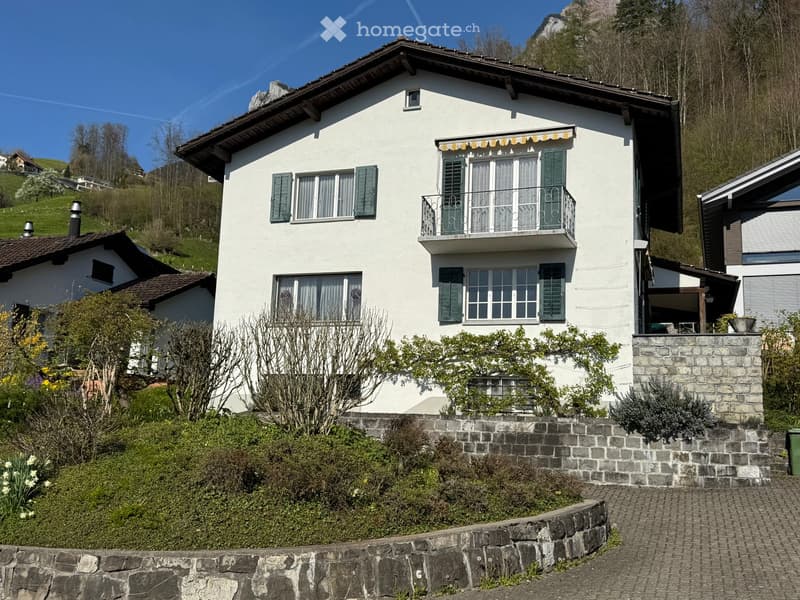 2 story house, white walls, brown roof, green shutters, balcony on second floor, surrounded by greenery and stone walls, nearby mountain landscape