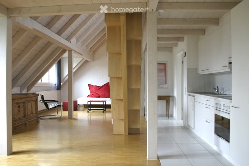 A wooden staircase, a white kitchen with white cabinets and tiles, a wooden table with a red pillow, a wooden chair, a bed, and a window.
