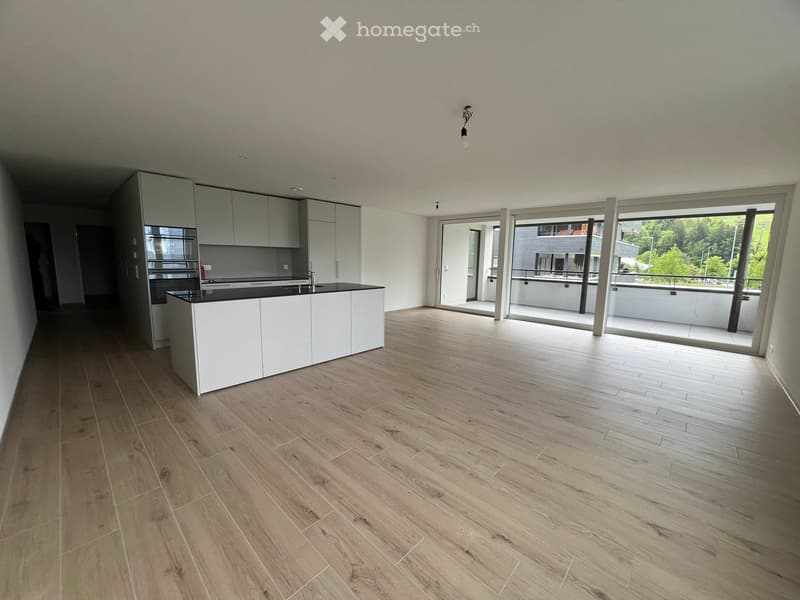 Empty kitchen with white cabinets, black countertop, and wooden flooring, leading to a balcony area with a view of greenery.