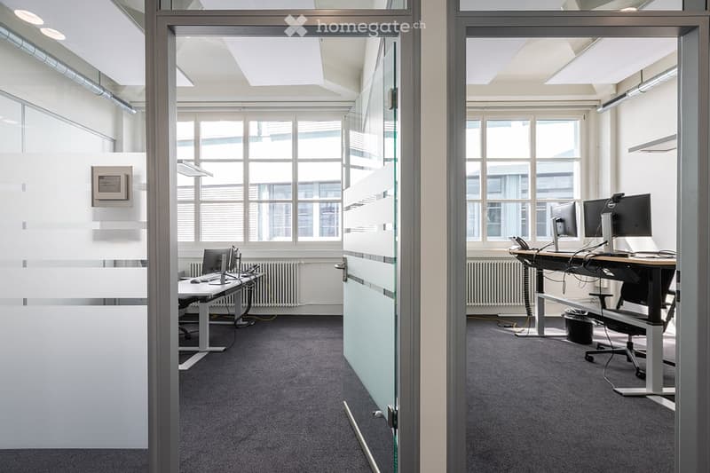 Office space with desks, chairs, monitors, radiators, carpeted floors, windows with blinds, a trash can, and lights on the ceiling.