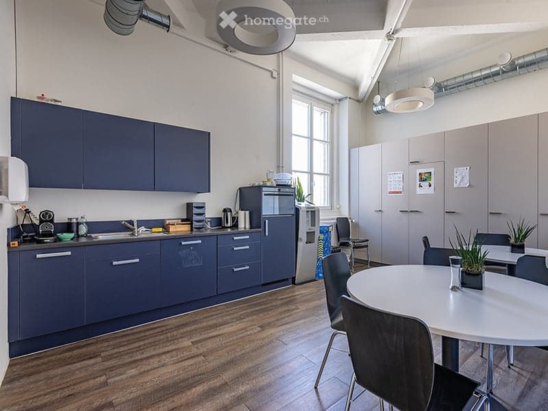 A modern, open-plan kitchen featuring blue cabinetry, stainless steel appliances, a wooden floor, and a glass window providing ample natural light.