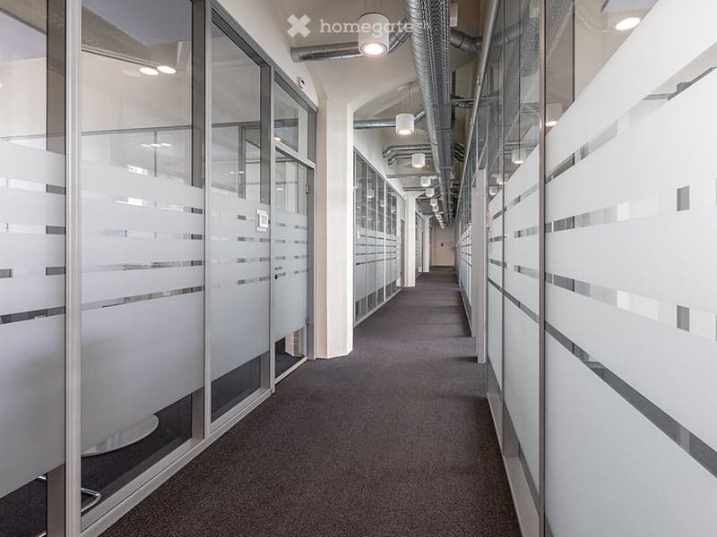 Office hallway with glass walls, metal ceiling pipes, and tiled floor.
