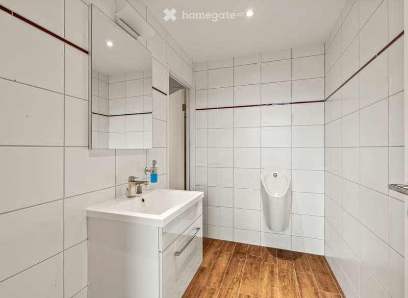 modern and clean bathroom with white tiled walls, wooden floor, a white vanity with a sink and urinal, and a mirror