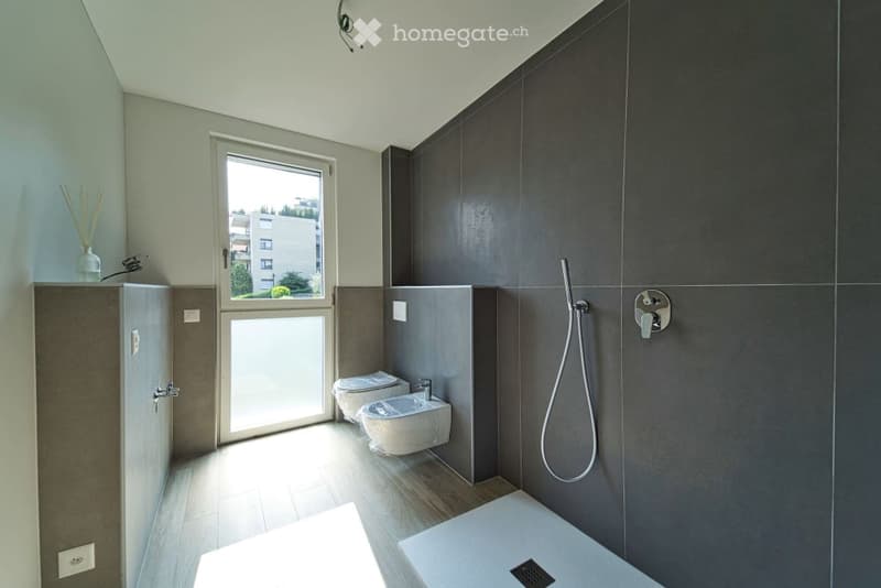 A contemporary bathroom with floor-to-ceiling tiles, glass window, double washbasin, two flush toilets, shower area with showerhead, and a small gray wall cabinet.