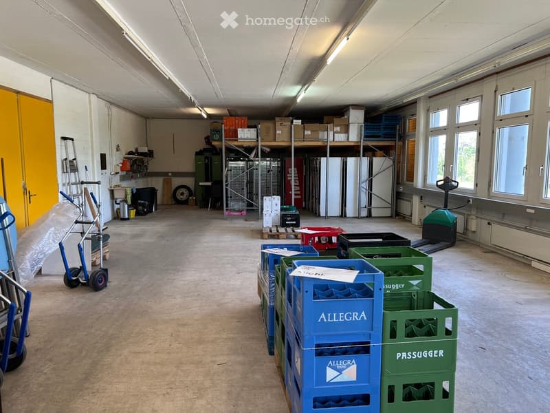 Large, empty warehouse space with concrete floors, white walls, and large windows. Shelves, crates, and storage items are present but no furniture.