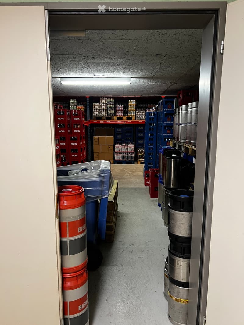 Storage area with concrete walls and floor, shelves holding beer kegs, crates, and boxes, illuminated by overhead lighting.