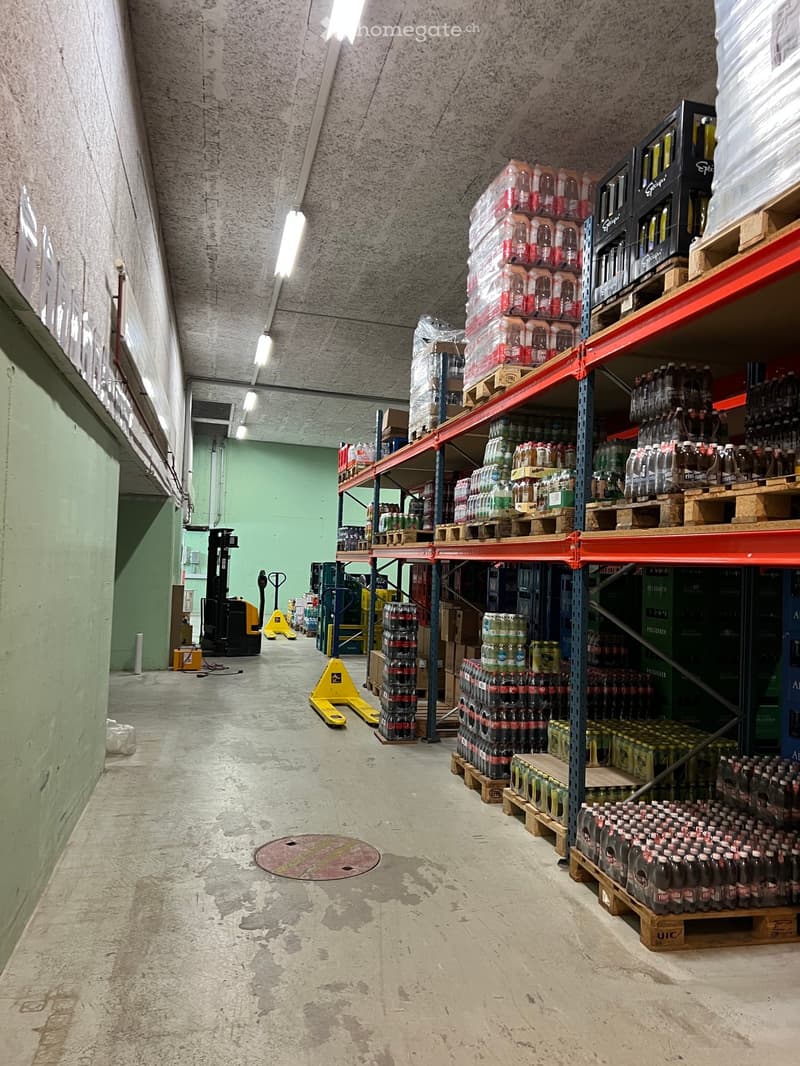 Warehouse with metal shelving units filled with bottled drinks on pallets, forklift in the background, concrete floor, exposed ceiling with fluorescent lighting.