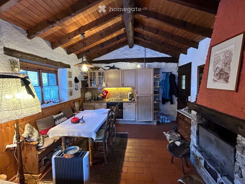 Wooden table and chairs, white tablecloth, lamp, fireplace, kitchen area with cabinets, countertop, and stove, window with blue curtains, wooden flooring, wooden beam ceiling