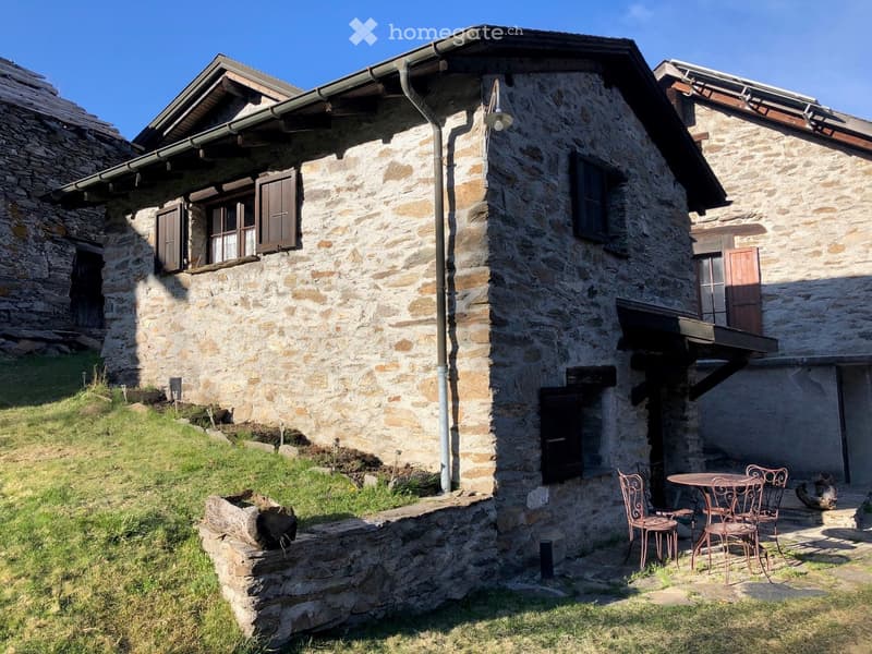 Stone wall house with wooden windows and doors, outdoor table with chairs, located on a sloped grassy terrain