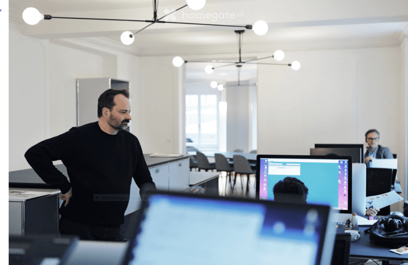 open office layout, white walls, ceiling lights, computer setup, desk, chair, man standing