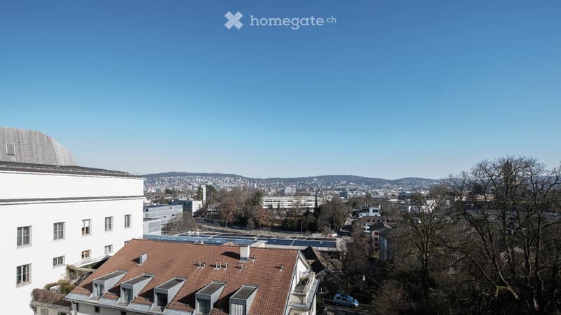 Aerial view of city, buildings, green trees, solar panels on the roof, mountains in the distance, clear blue sky