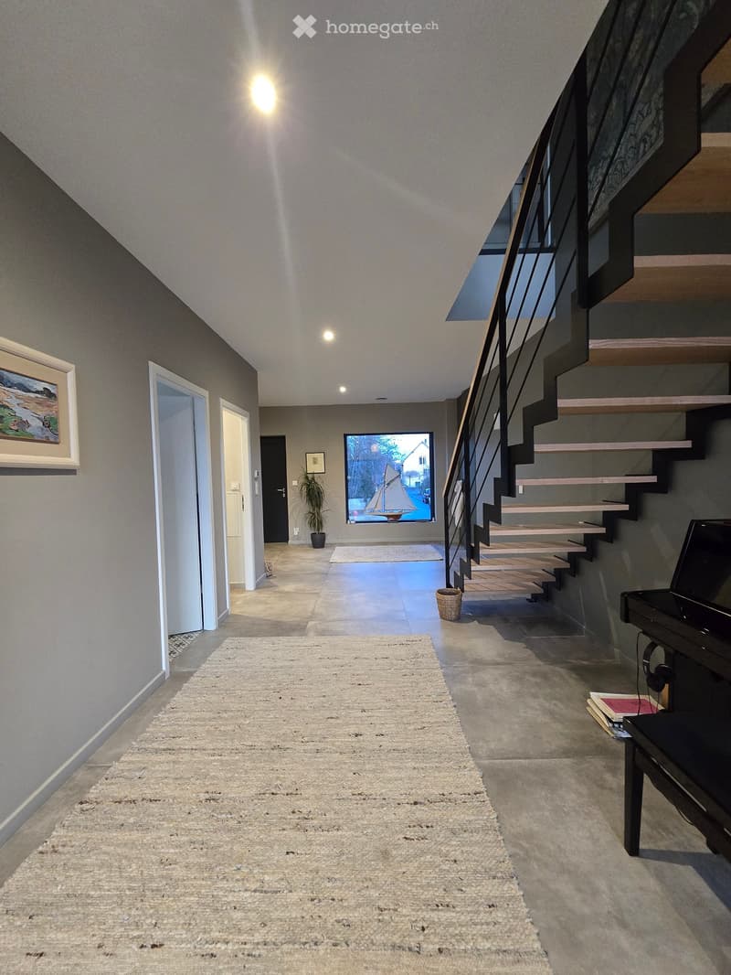 large open hallway, wooden staircase, grey carpet, concrete floor, entrance to multiple rooms, large window with sailboat view, plant, piano