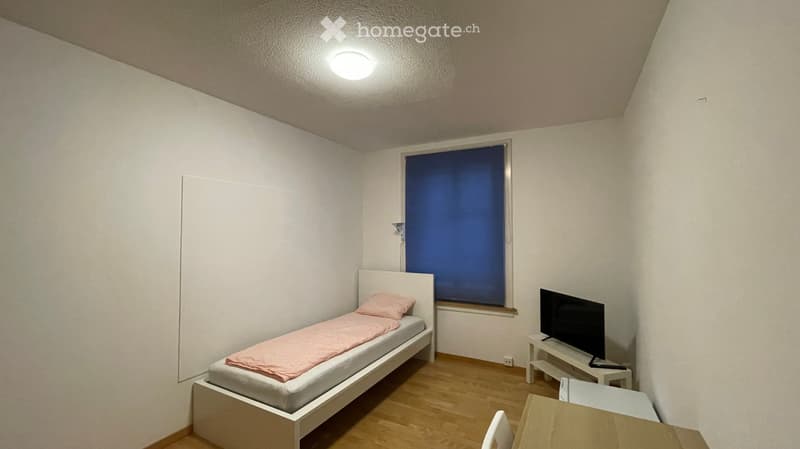 A room with white walls, a bed, wooden floor, a TV on a stand, and a window with blue curtains.