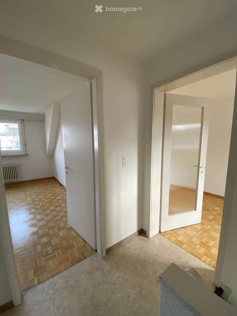 Empty room with white walls, wooden floors, and white doors, one with a glass panel, a radiator on the wall, and a window with an outside view.