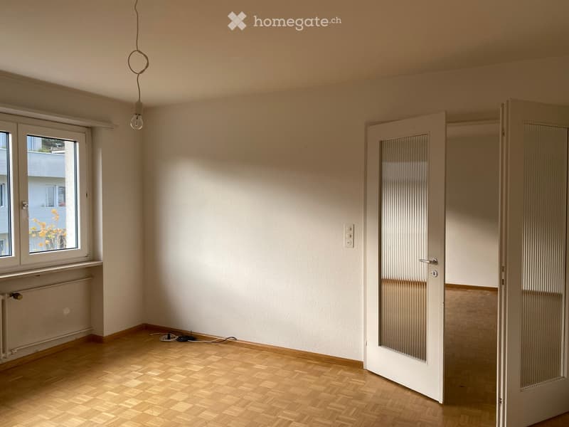 Empty room with white walls, wooden floor, window with outdoor view, radiator, doors