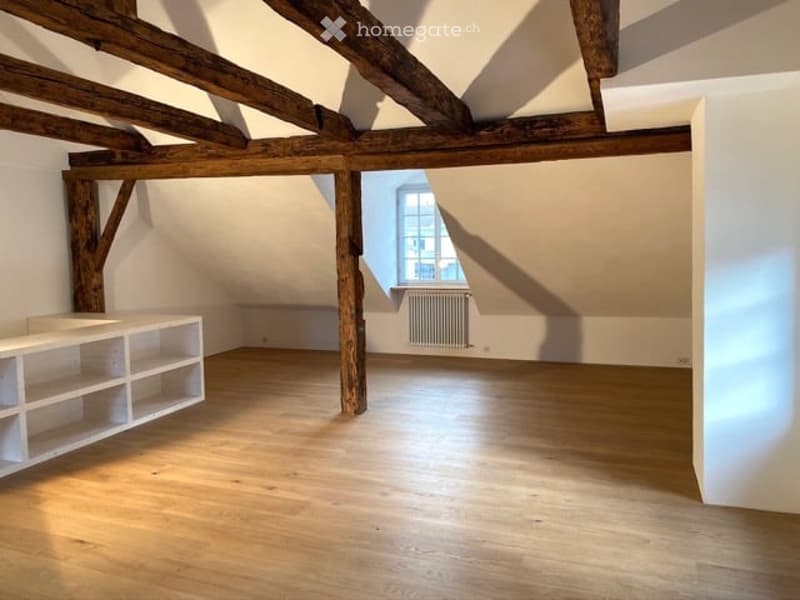 Empty room with wooden floor and exposed wooden ceiling beams, small white shelf, white wall, small window with white frame.