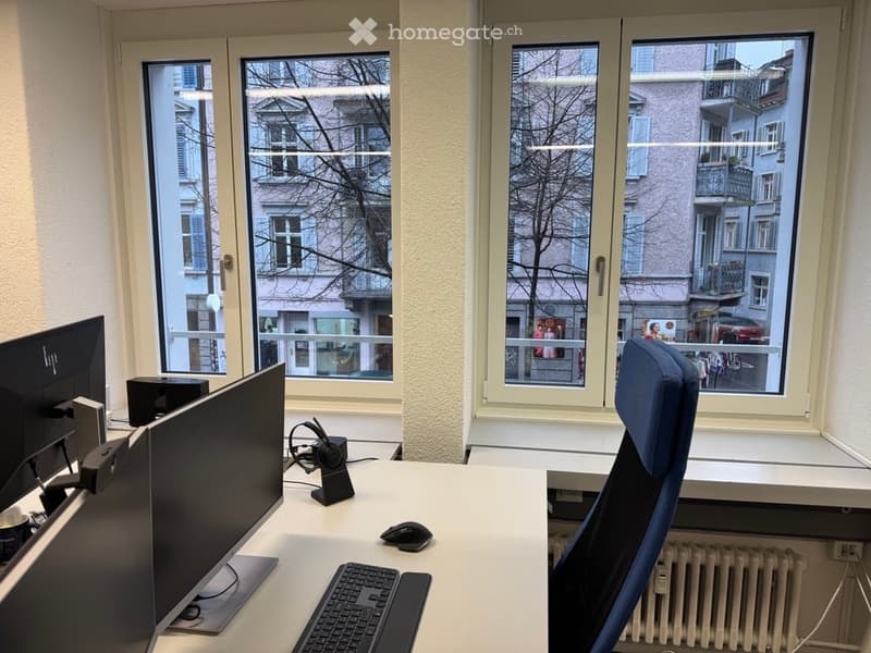 Office space, two computer monitors, keyboard, mouse, blue chair, double windows, white desk, two large windows facing street view