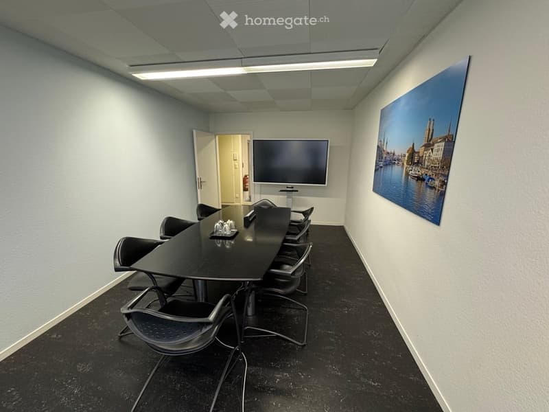 empty meeting room with long table, 13 chairs, TV mounted on wall, picture frame on right wall, exit door on left wall