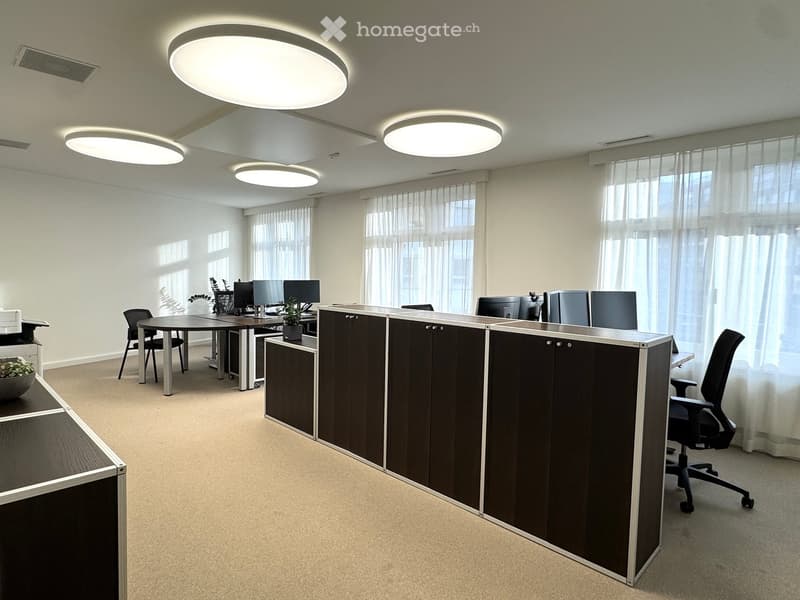 Modern office interior with beige carpet, beige walls, and white ceiling, equipped with a large desk, computer setup, office chairs, round table, a black cabinet, and multiple round ceiling lights.