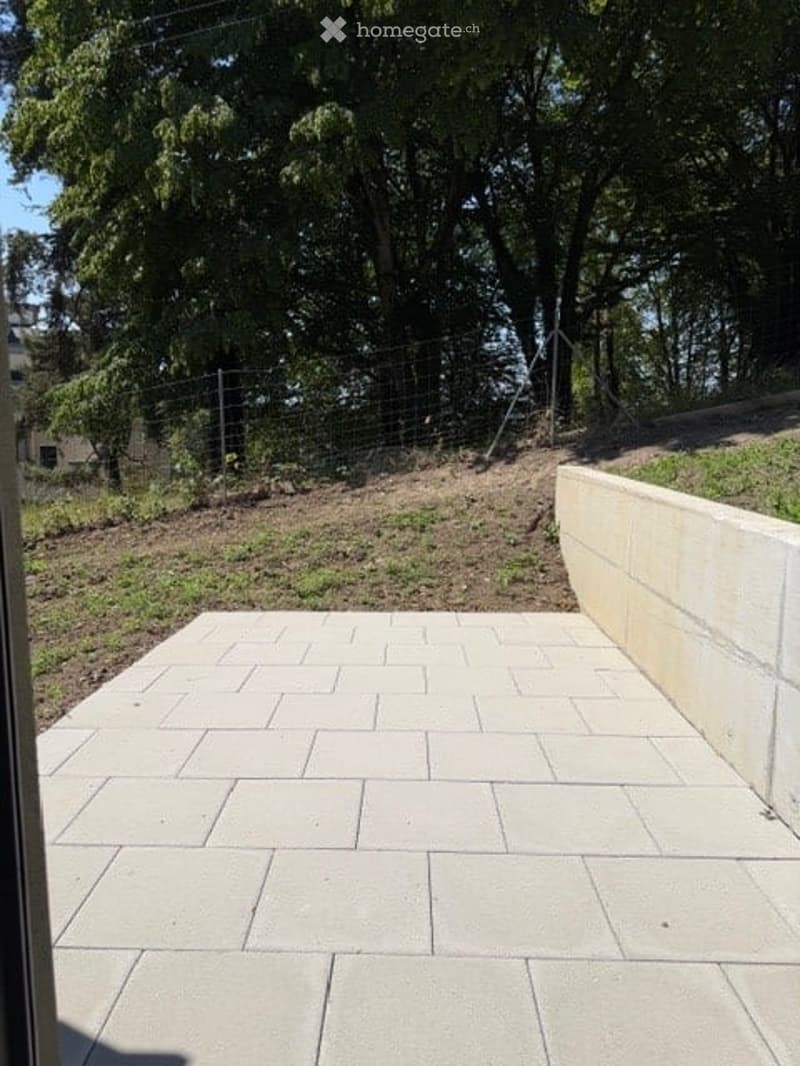 Outdoor patio with light-colored stone tiles, surrounded by a low concrete retaining wall and a wire fence, with trees and greenery in the background.