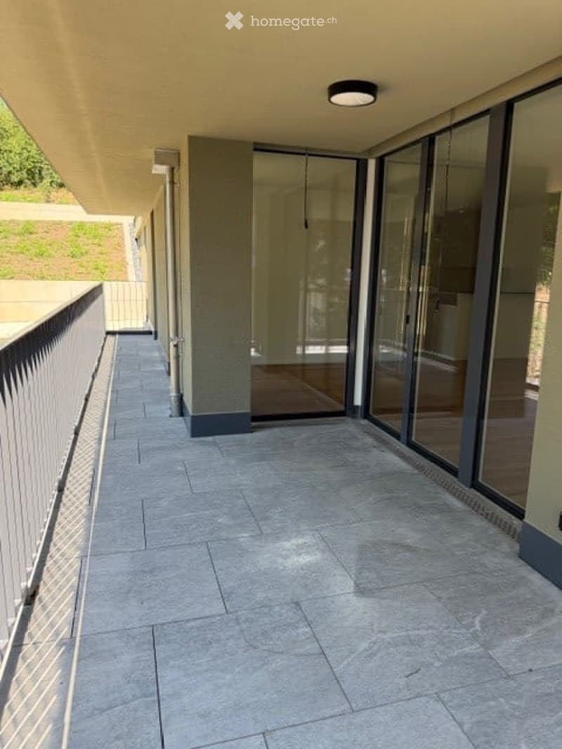 Modern balcony with grey tiled flooring, white metal railing, large glass sliding doors leading to interior, recessed ceiling light, and a view of greenery beyond.