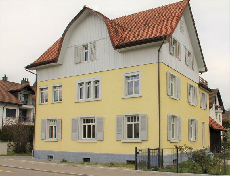 Yellow painted house with red tiled roof, multiple windows with white shutters, raised ground floor, black gates in front of house, green shrubs in front