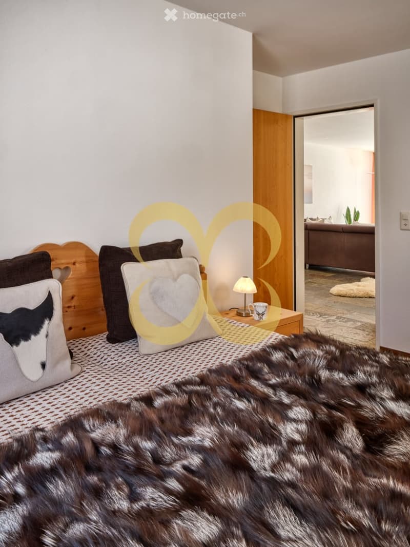 Bed with a cow head pillow, a bedside table, wooden headboard, wooden floor, white walls, natural light