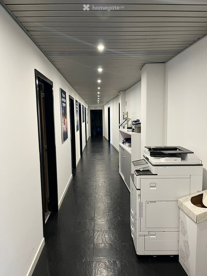 Long corridor with black tiles and white walls, closed doors along the sides, two printers on the right, a storage unit with a display in the distance, and some illuminated lights on the ceiling.