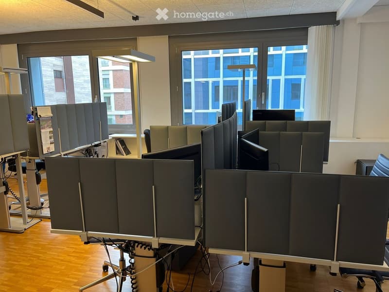 Office room with partition screens, multiple computer monitors, white flooring, large windows, office furniture, two floor lamps, white wall.