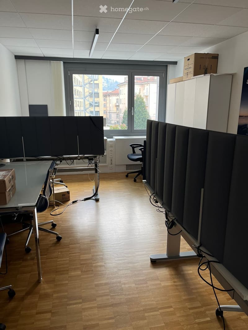 An empty office with wooden floors, white walls, large windows, and adjustable desks. Chairs, a desk with monitors, and storage boxes are present. A lamp is mounted on the ceiling.