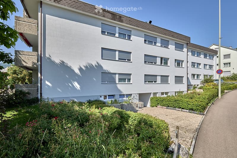Apartment building with white exterior, multiple balconies, windows with shutters, small gardens in front